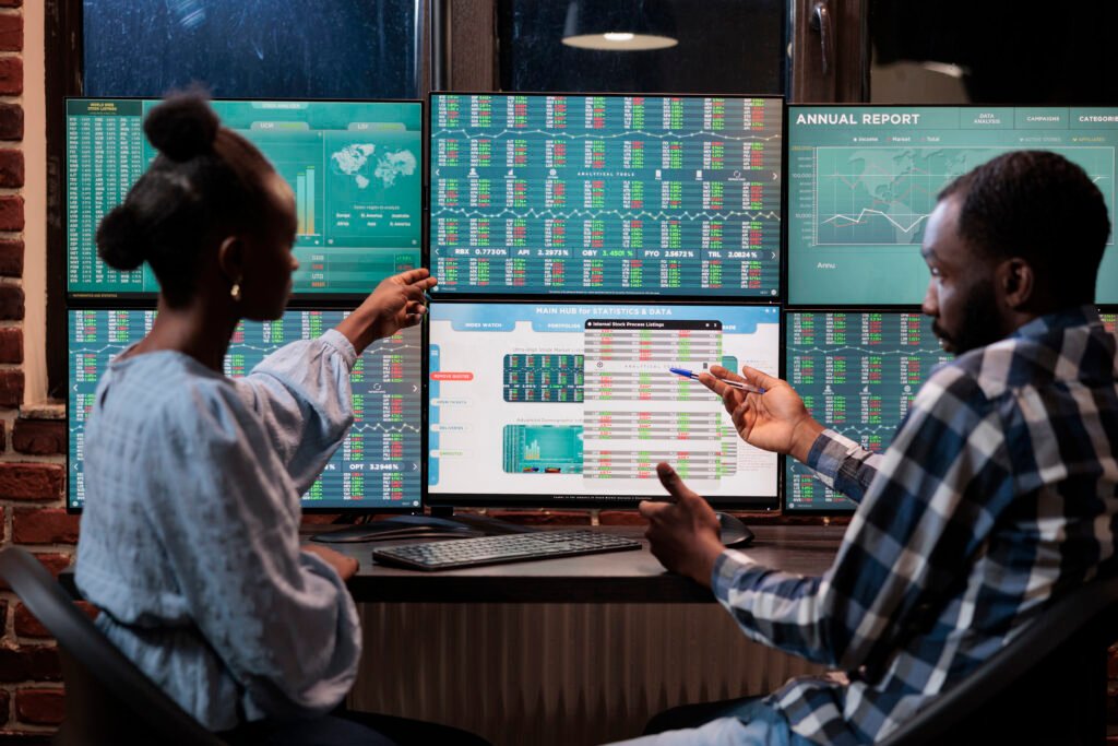 Two Black fintech professionals, a woman with a bun and a man in plaid, point at stock charts and annual reports on multiple monitors in a dimly lit office.