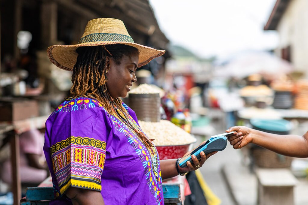 A smiling African woman in vibrant purple traditional attire with colorful patterns and a wide straw hat stands at an outdoor market stall. She holds a blue mobile payment terminal while accepting a card from a customer's hand, surrounded by market goods like grains and baskets in a bustling rural or semi-urban setting.