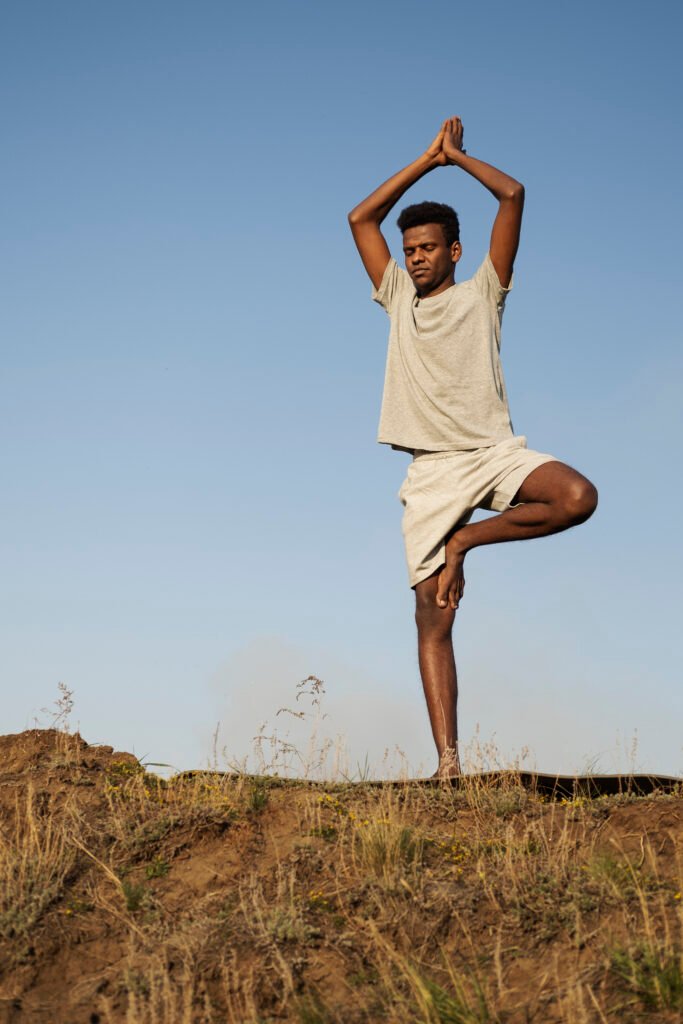 équilibre,: Un jeune homme africain effectuant la posture de l'arbre en yoga en plein air sous un ciel bleu clair ; représentant l'équilibre physique, la santé mentale et l'importance du bien-être au quotidien.